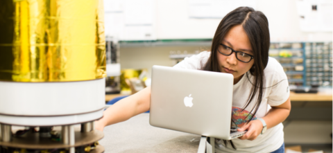 Woman holding laptop and reaching for cylinder in lab