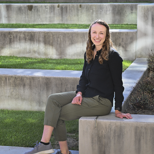 Portrait of Christine in a black top sitting alongside cement steps and grass. 